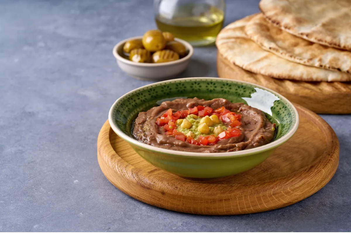 Ful medames in a green bowl topped with diced tomatoes, chickpeas and herbs, with olives, olive oil and flatbread behind it.