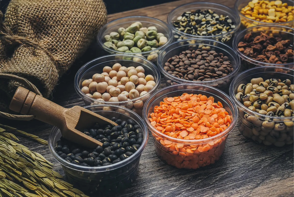 Bowls of dried chickpeas, red and brown lentils, black beans, green peas and black-eyed peas on a wooden table, beside a burlap sack and a wooden scoop.
