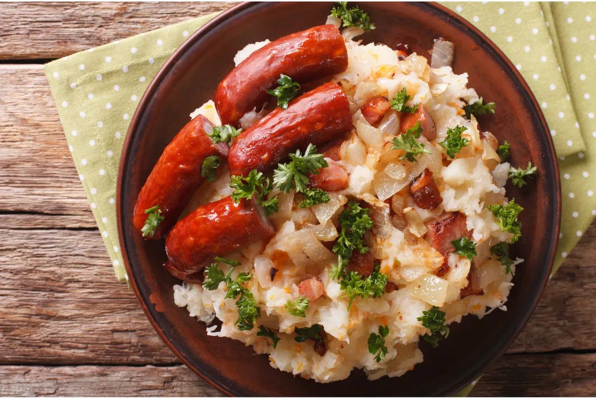 A plate of stamppot: mashed potatoes with onions and bacon, topped with smoked sausages and parsley.